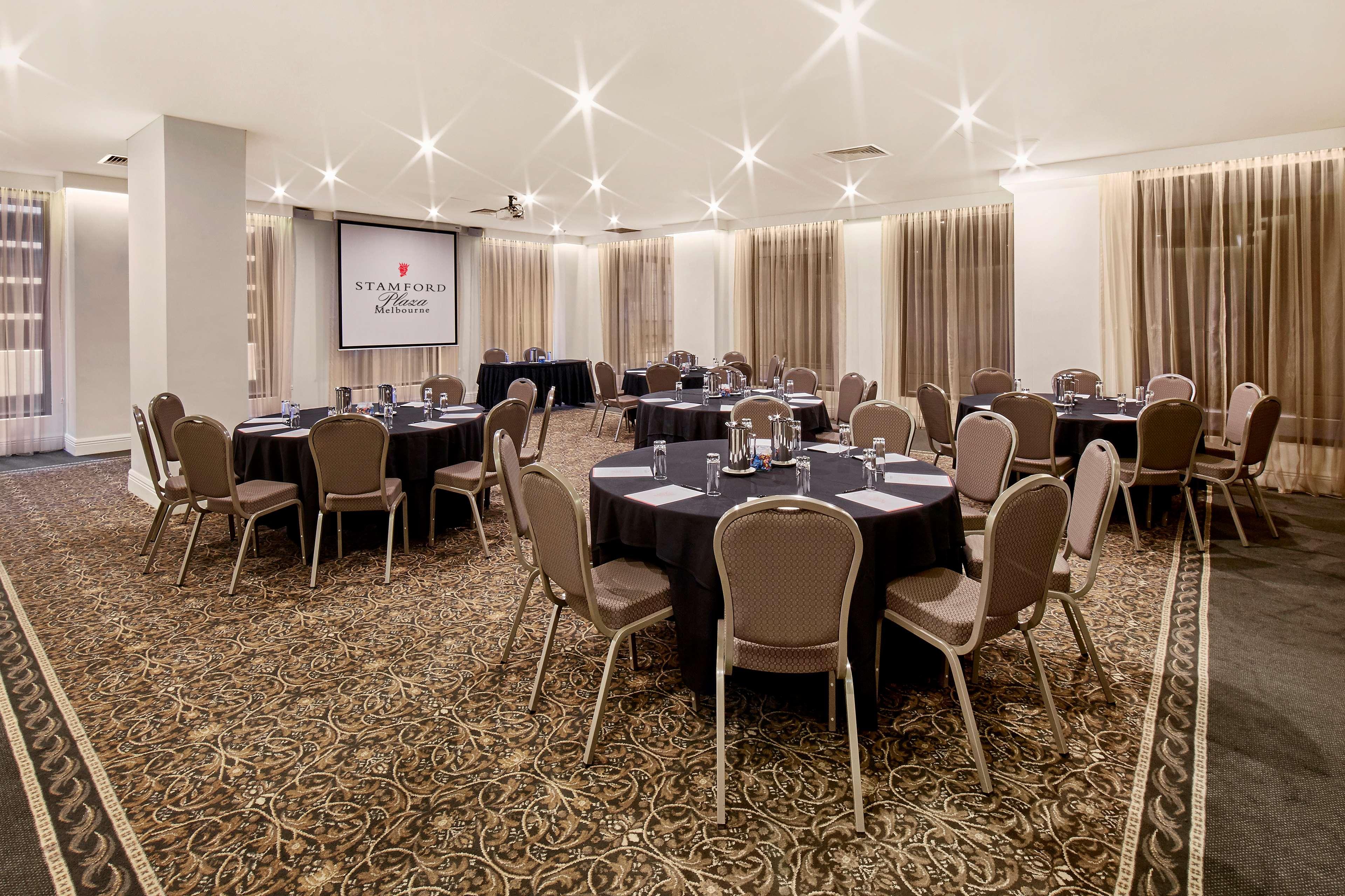 Hotel Stamford Plaza Melbourne Esterno foto The photo shows a well-lit event space set up for a gathering or meeting. There are several round tables covered with black tablecloths, each surrounded by beige chairs. In the background, a large screen displays a logo or branding, likely related to