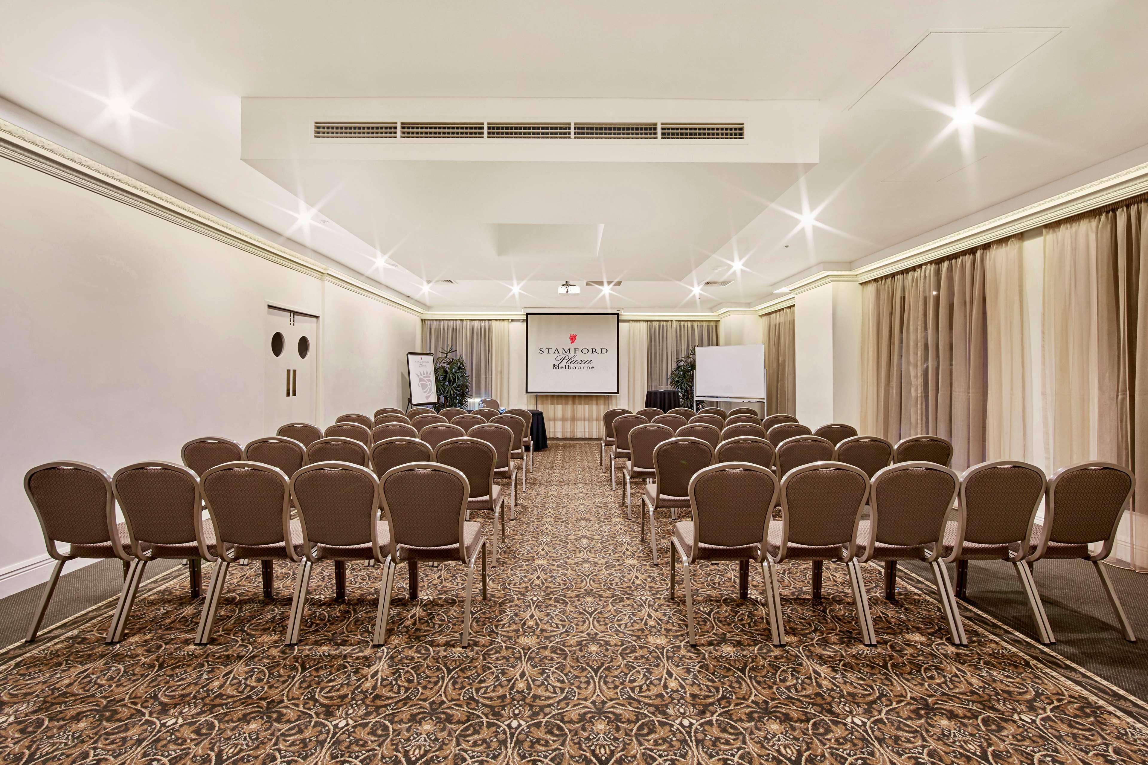 Hotel Stamford Plaza Melbourne Esterno foto The photo shows a conference room set up for an event. It features several rows of beige chairs arranged facing a central presentation area. At the front, there is a screen displaying a logo or title, possibly for a conference or meeting. The walls h