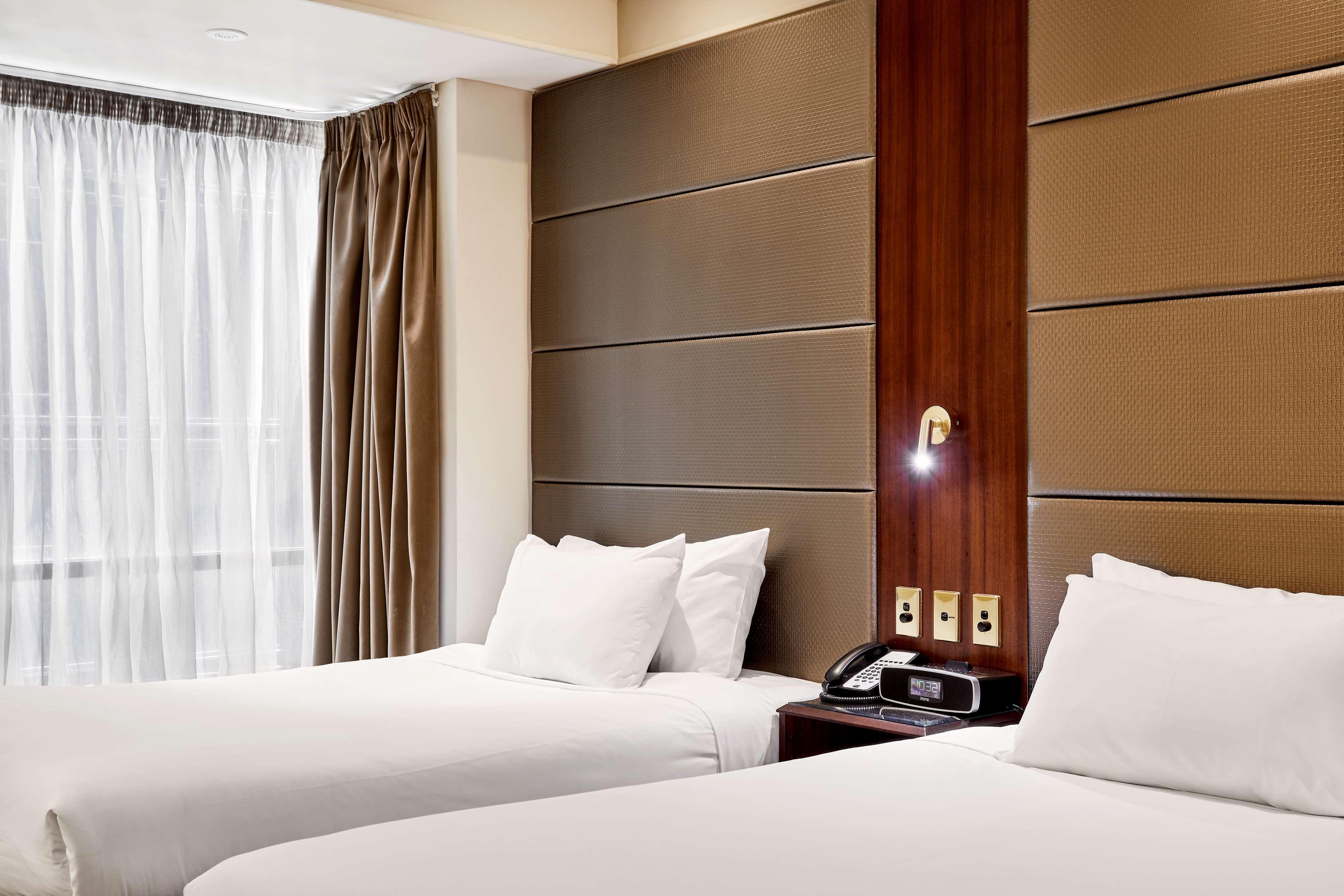 Hotel Stamford Plaza Melbourne Esterno foto The photo shows a modern hotel room featuring two twin beds with white linens and fluffy pillows. The walls behind the beds have a combination of textured panels in neutral colors. There is a bedside table with a lamp and a phone. A large window with