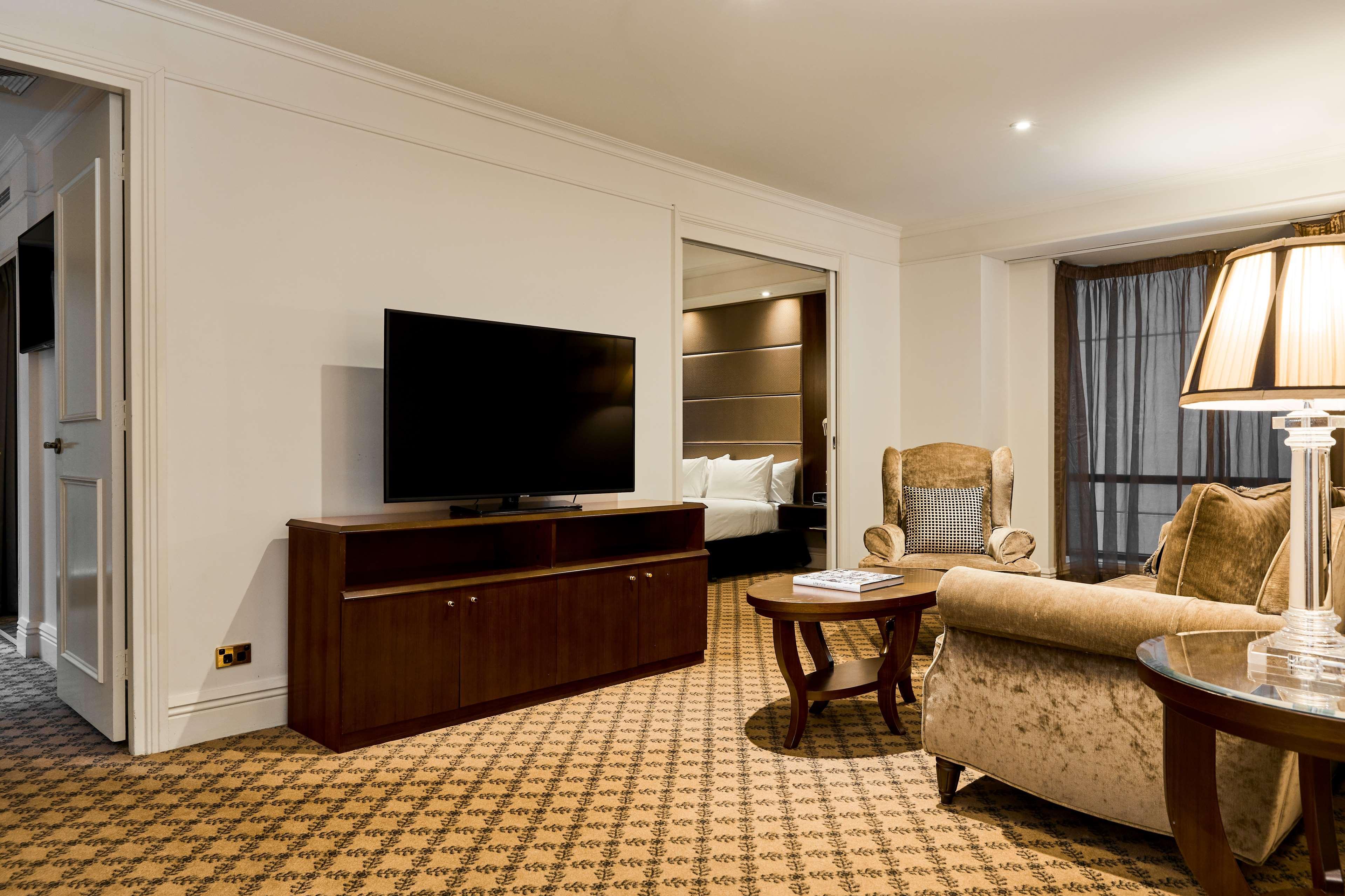 Hotel Stamford Plaza Melbourne Esterno foto The photo shows a well-furnished room, likely a hotel suite or a living area. There is a large flat-screen television mounted on a wooden entertainment unit. In front of the TV, there is a round wooden coffee table. To the side, a comfortable armchai