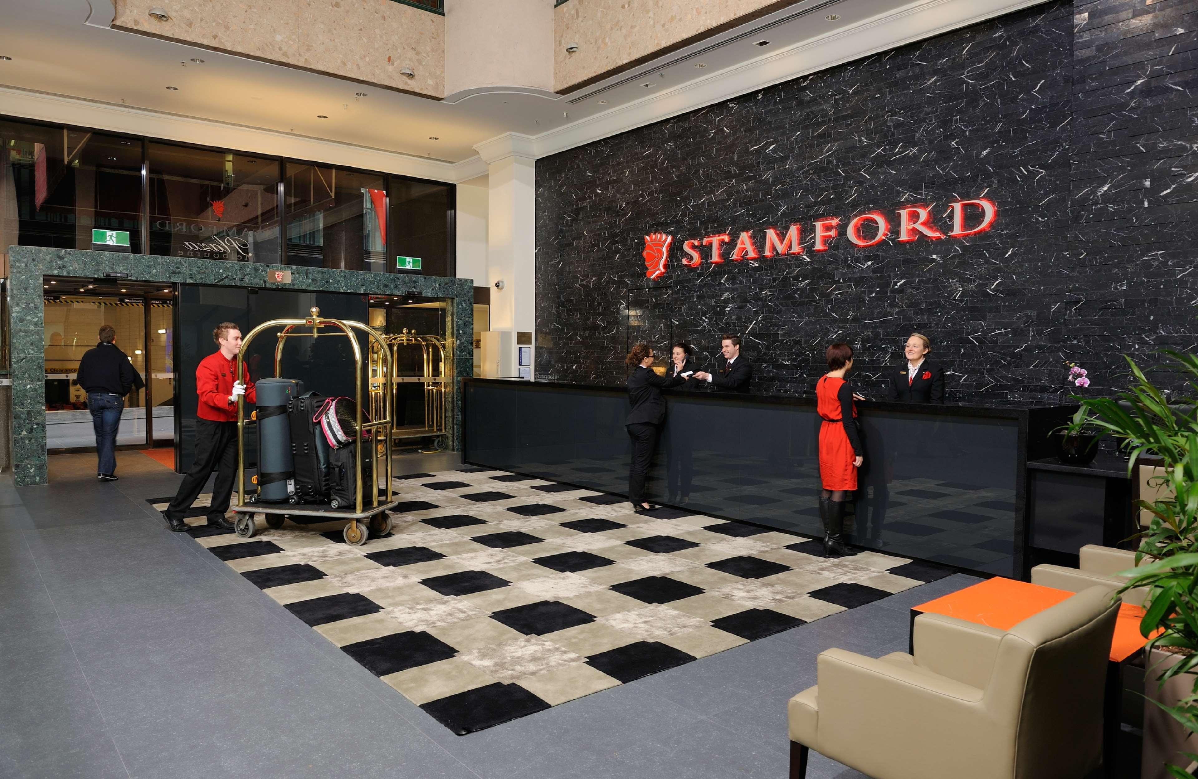 Hotel Stamford Plaza Melbourne Interno foto The photo shows the interior of a hotel lobby, specifically the Stamford Hotel. There is a large reception desk where several staff members are attending to guests. In the foreground, a bell cart loaded with luggage is positioned beside the reception
