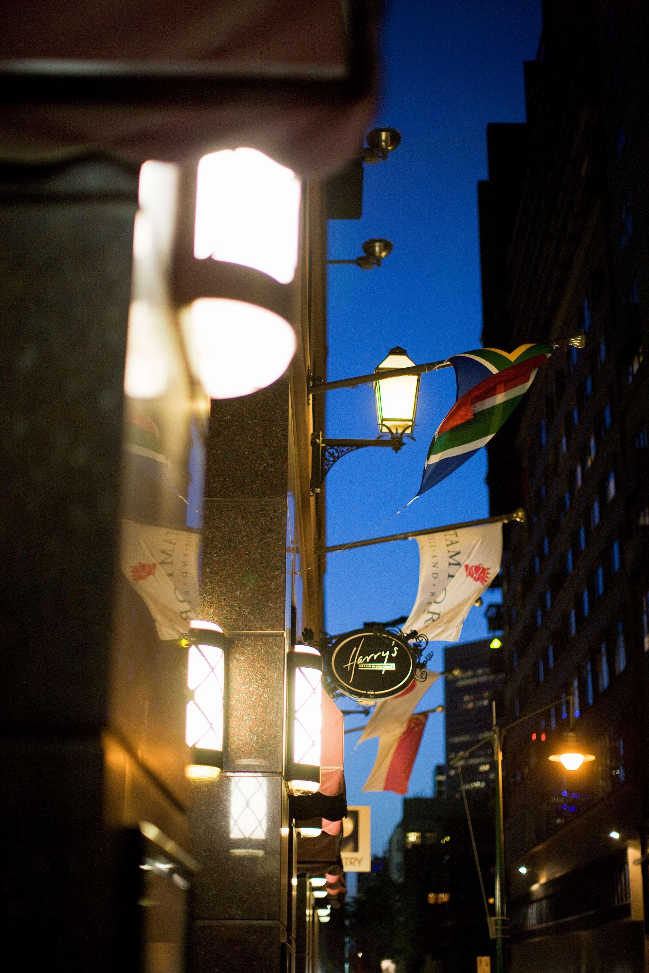 Hotel Stamford Plaza Melbourne Esterno foto The photo features a street view of a building at dusk. Prominently displayed are several flags, including a South African flag, which are fluttering in the wind. There is a sign that reads "Harry's," likely indicating the name of a business or estab