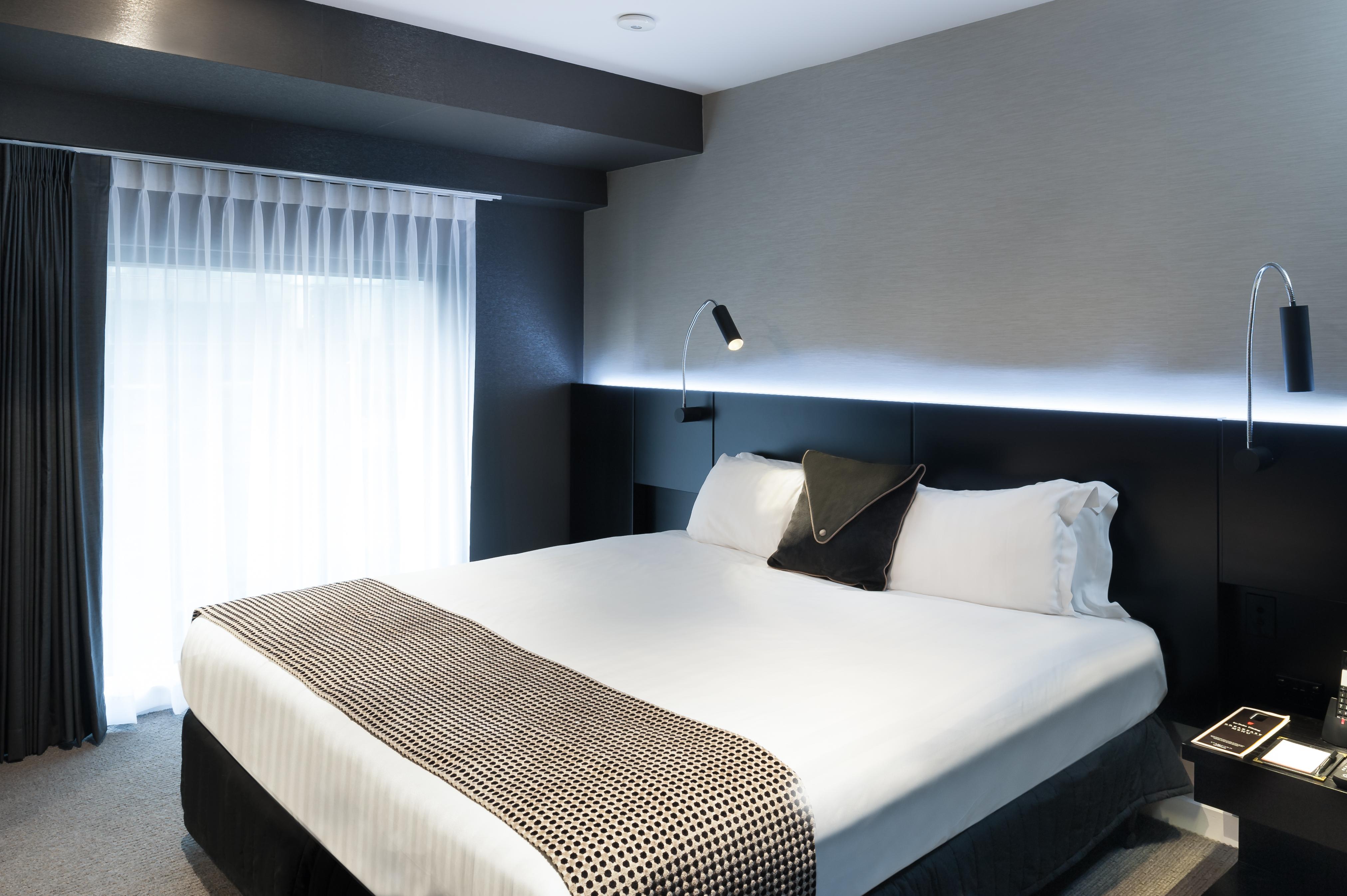 Hotel Stamford Plaza Melbourne Esterno foto The photo shows a modern hotel room featuring a large bed with white linens and a decorative throw blanket. The room has a sleek and contemporary design, with gray walls and a soft lighting strip along the headboard. A window is adorned with sheer cu