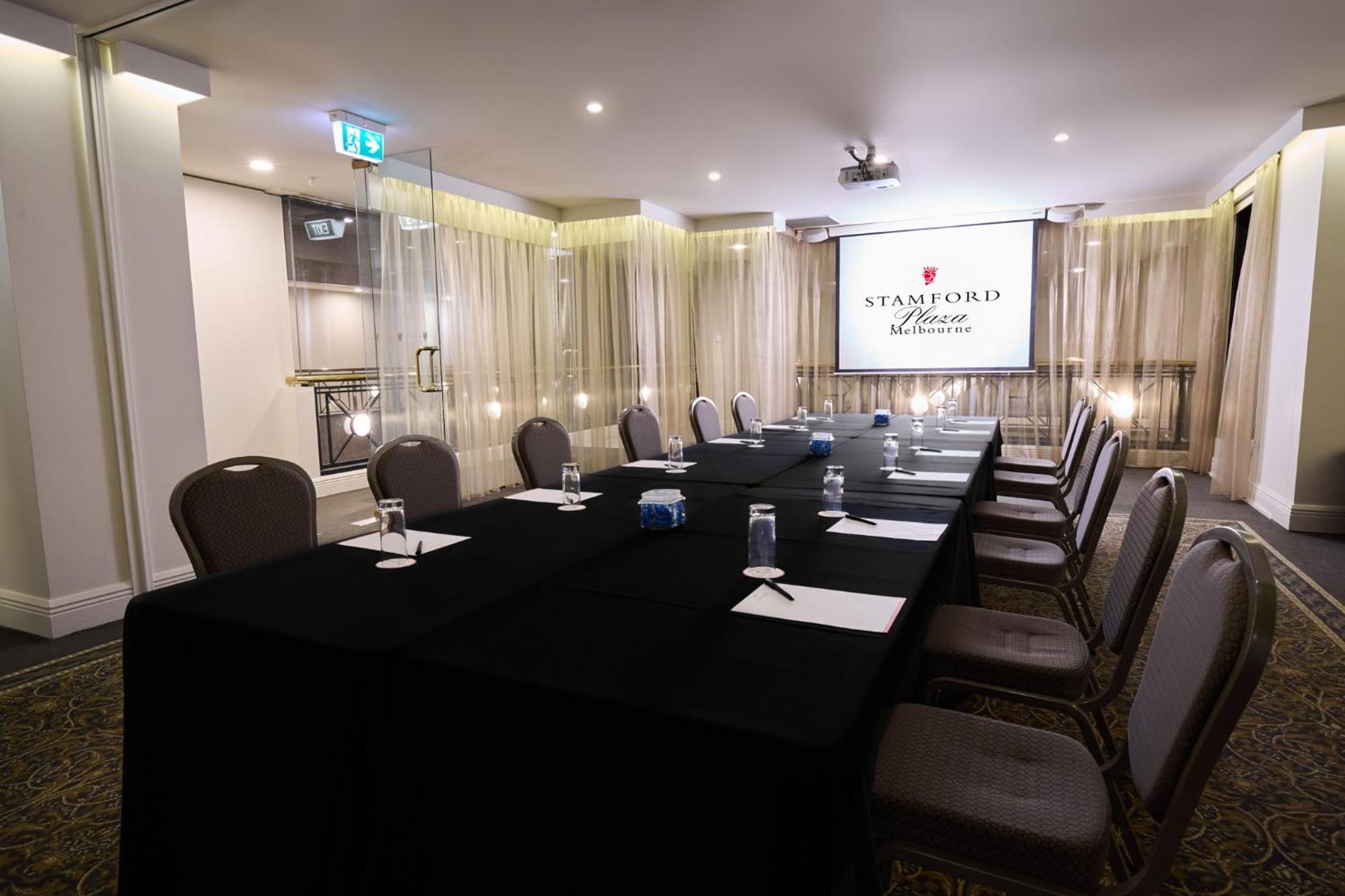 Hotel Stamford Plaza Melbourne Esterno foto The photo shows a well-appointed conference room designed for meetings or presentations. There is a long rectangular table covered with a black tablecloth, surrounded by upholstered chairs. On the table, there are several folded pieces of paper and g
