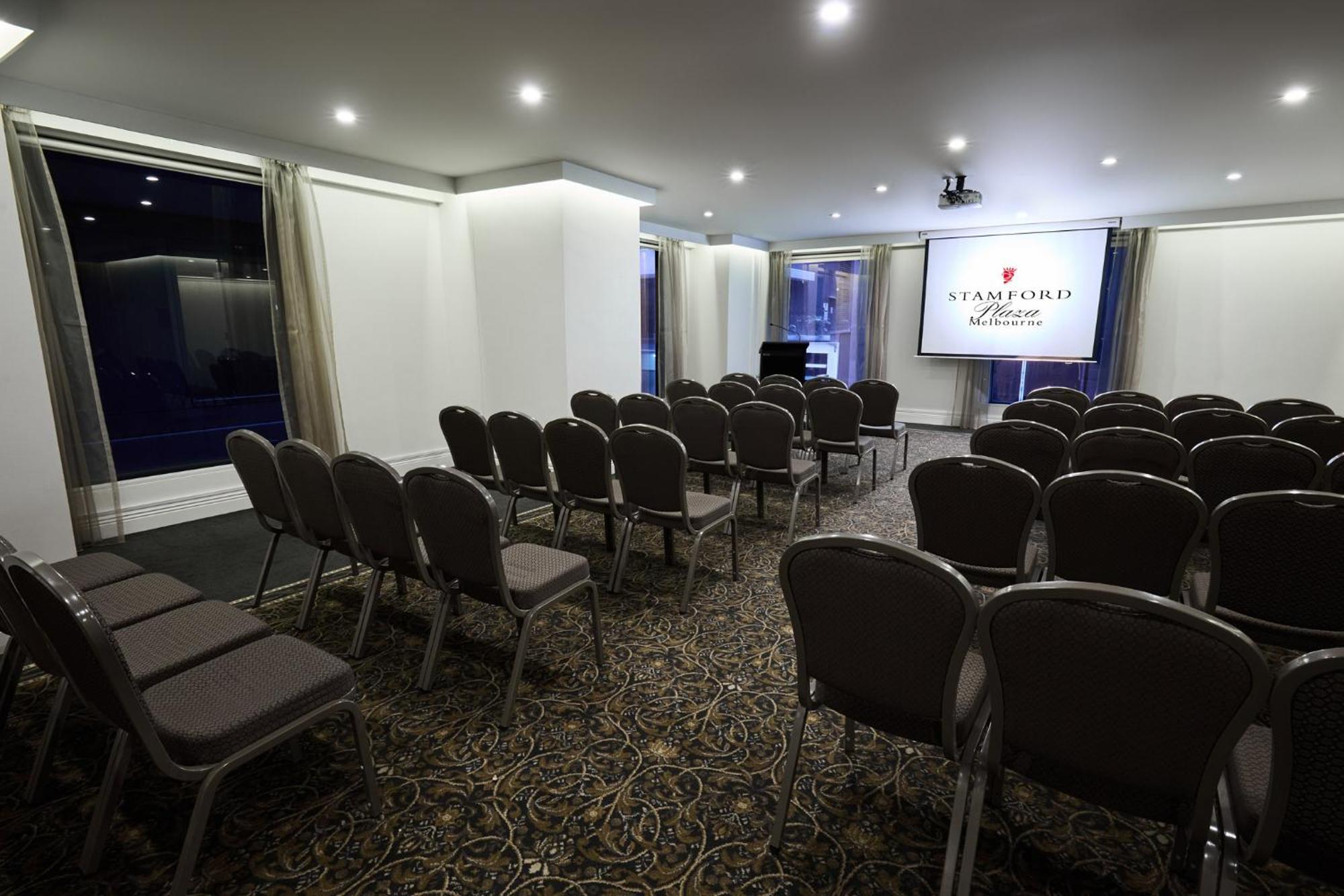 Hotel Stamford Plaza Melbourne Esterno foto The image shows a conference or meeting room set up for an event. There are rows of gray chairs arranged facing a large screen displaying the name "Warford House." The room is well-lit with overhead lighting and has large windows covered with light-c